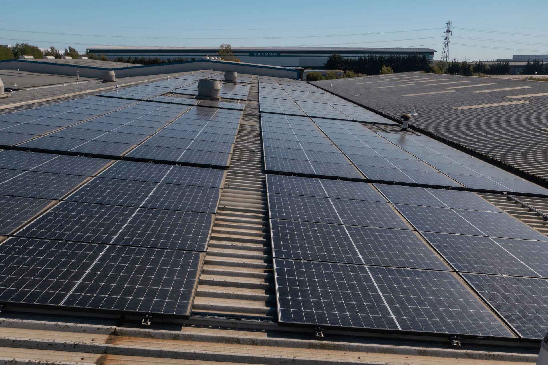 Rows of solar panels installed on a flat industrial roof, extending toward a distant building under a clear blue sky.